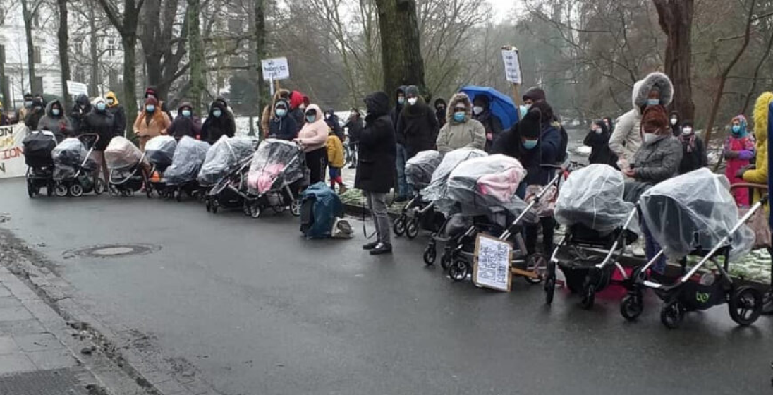 A group of protestors demonstrating in a town square, holding banners and strollers as protest symbols.