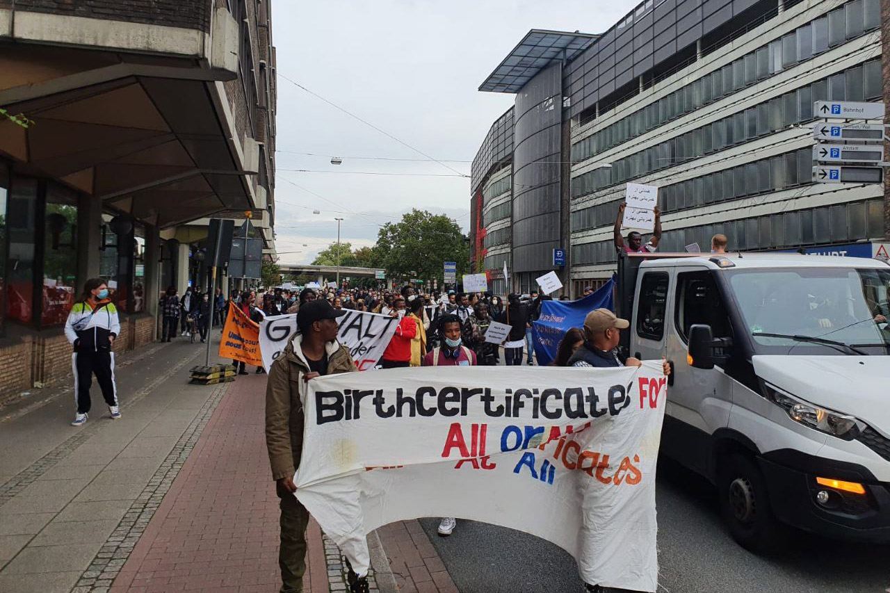 Protestors marching down a street and carrying a banner that reads "birth certificates for all or no birth certificates at all."